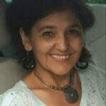 O femeie cu păr negru și cercei zâmbește camerei. Ea poartă un top de dantelă de culoare deschisă și un colier cu un pandantiv rotund. Fundalul pare să fie interiorul unei mașini.